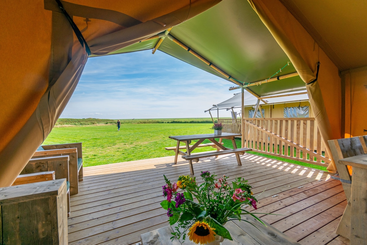 An open view from a safari tent showcases a spacious deck with a wooden table and benches. A bouquet of flowers sits at the center, while the grassy field extends towards the horizon. The entrance reveals bright skies and a peaceful landscape.