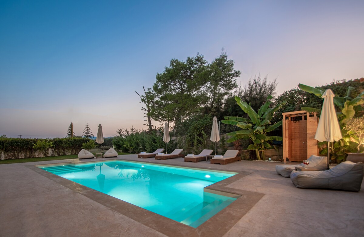 An inviting swimming pool is illuminated with a soft blue glow, surrounded by comfortable sunbeds. Lush greenery and a few tall trees are visible in the background, while umbrella shades provide shelter from the sun. A cozy seating area is set beside the pool.