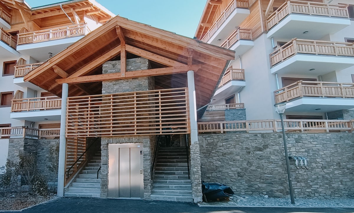 A modern building entrance features a wooden gabled roof supported by stone pillars. An outdoor elevator is visible, surrounded by natural stone walls. Balconies with wooden railings extend from the upper floors, allowing for outdoor views. A pathway leads to the stairway, blending with the entrance.