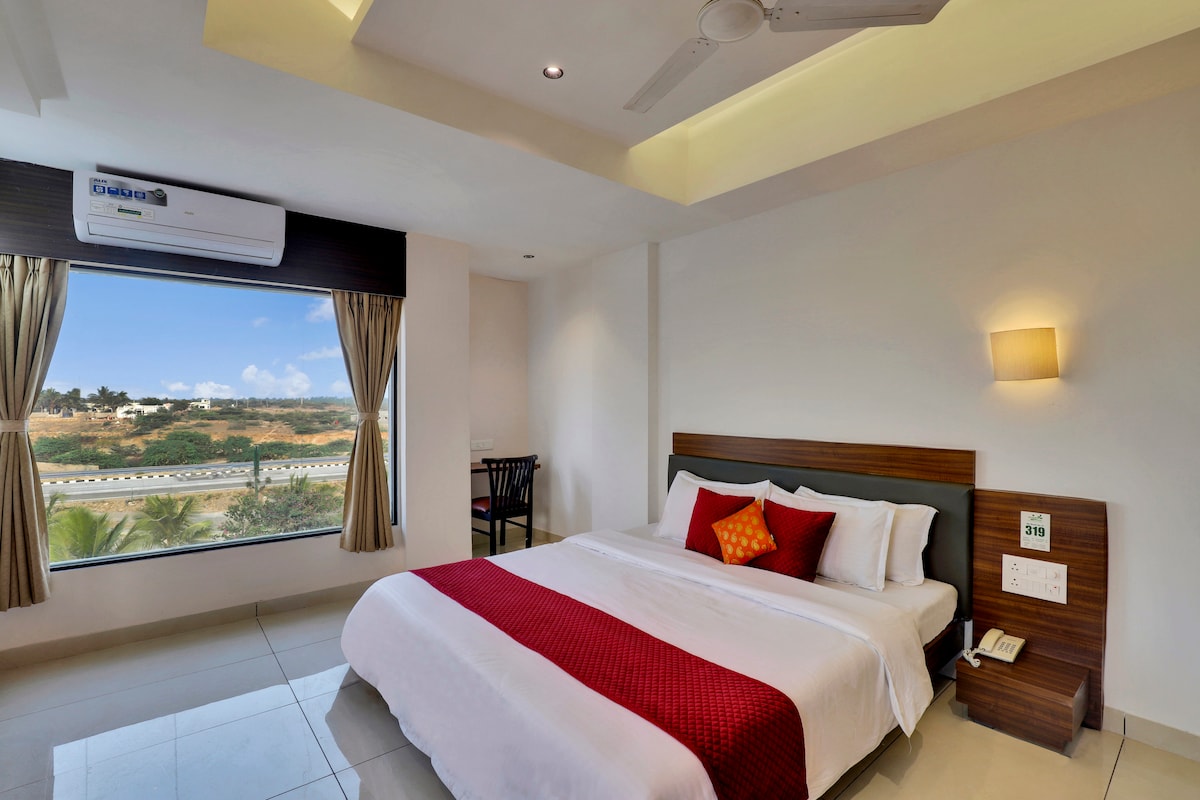 A bright family room features a spacious bed with a red accent throw pillow, complemented by light-colored linens. Large windows provide a view of lush greenery outside, while a small dining table is positioned nearby. Soft lighting and modern decor create a welcoming ambiance.