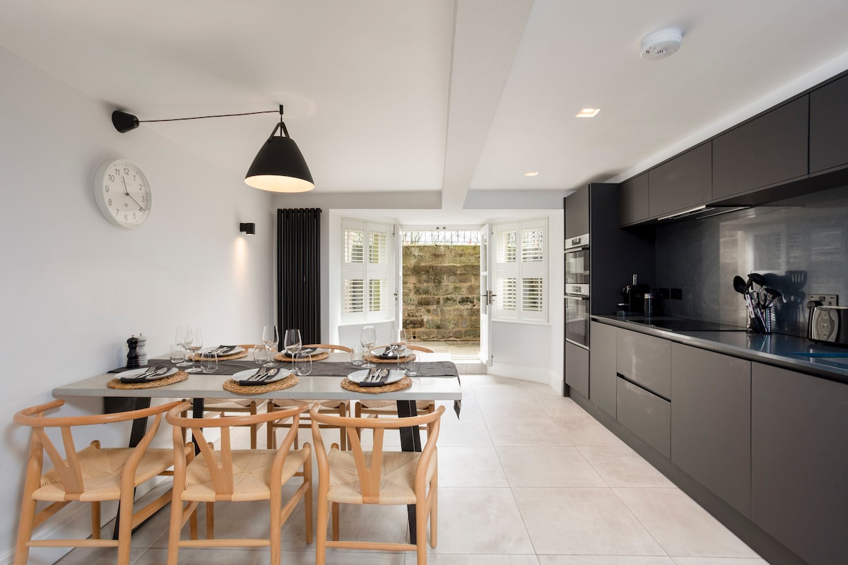A contemporary kitchen and dining area features sleek dark cabinetry paired with modern appliances. A wooden dining table is set for six, accompanied by stylish chairs. Light enters through large windows, illuminating the space and revealing a view of a stone wall beyond.