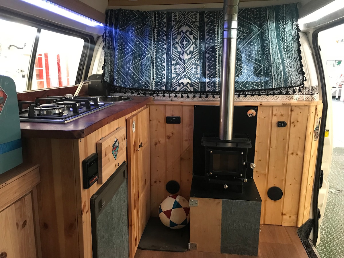 The interior of the van features wooden paneling with a compact kitchen area. A gas stove is positioned above a small sink, while a cozy fireplace stands against the wooden wall. Colorful fabric hangs above the cooking area, adding a touch of warmth.