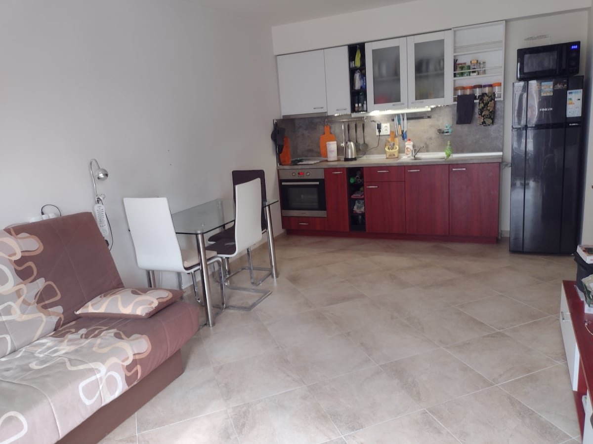 The living area features a foldable sofa, a dining table with four chairs, and a well-equipped kitchen. Kitchen appliances including a refrigerator, oven, and storage cabinets are visible. Light-colored tiled flooring enhances the spacious and functional setup.