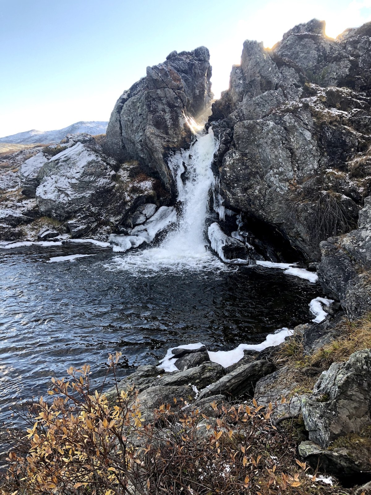 A waterfall cascades over a rocky formation, flowing into a tranquil body of water. Surrounding vegetation includes patches of grass and small shrubs, with some areas displaying evidence of early snow. The scene is set against a backdrop of distant mountains under a clear blue sky.