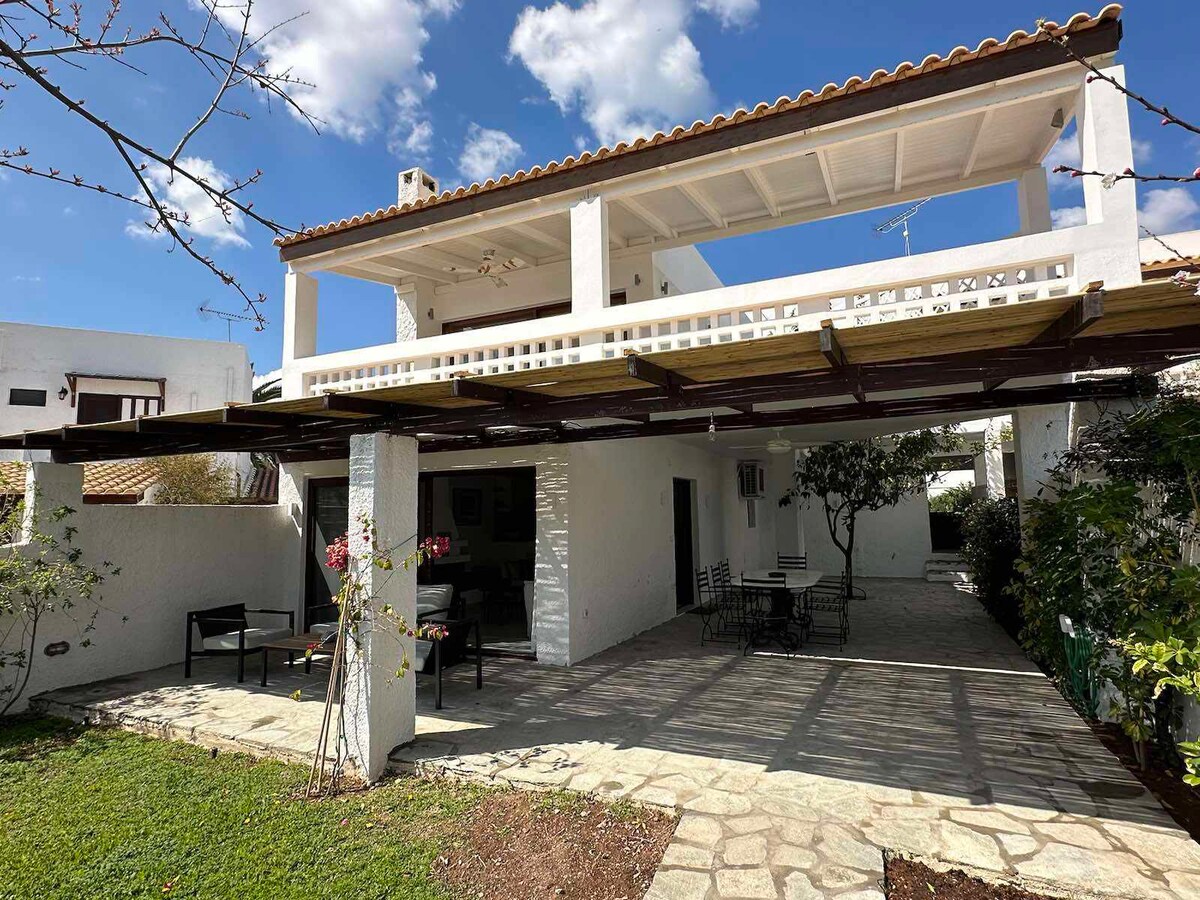 The exterior features a two-story white villa with a covered terrace area beneath a tiled roof. Stone pathways lead through the garden, where seating options are available. Sunlit clouds drift above, enhancing the serene outdoor environment.