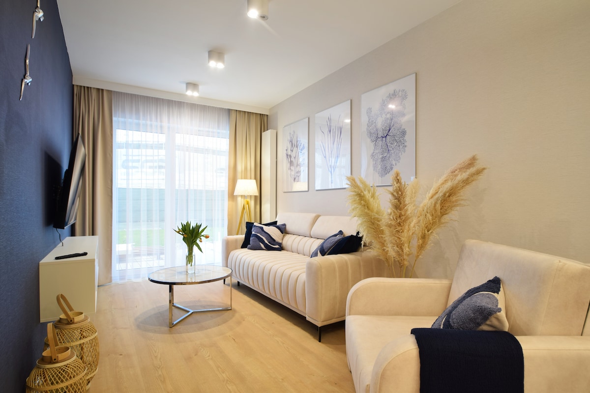 A light-filled living area features a comfortable sofa and an armchair in neutral tones. Decorative artwork adorns the walls, and a glass coffee table sits in front. A vase with greenery adds a touch of nature, while sheer curtains provide soft lighting.