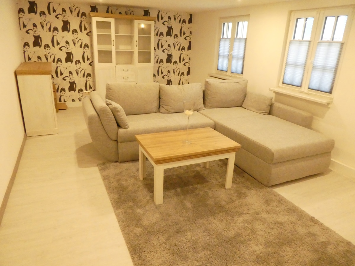 A comfortable living area is highlighted by a light gray sectional sofa, paired with a simple wooden coffee table. The wall features a subtle pattern, and natural light enters through two windows, enhancing the airy ambiance of the space.