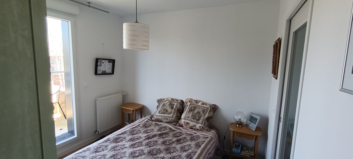 A serene bedroom features a spacious bed adorned with a patterned bedspread. Two bedside tables are positioned on either side, one holding a small lamp. Natural light pours in through a window, enhancing the bright, neutral walls.