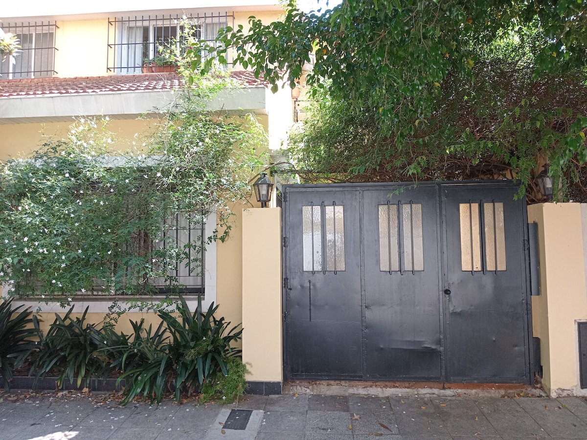 A black metal gate serves as the entrance to a residential property, bordered by lush greenery and low shrubbery. The gate features vertical slats, allowing glimpses of the inviting space beyond. Soft light filters through the foliage, creating a serene atmosphere.