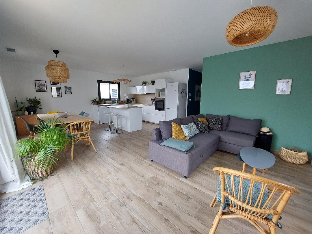 A spacious living area is shown, featuring a comfortable gray sofa adorned with colorful cushions. A round table with chairs is visible nearby, while the kitchen area is equipped with modern appliances. Natural light filters through large windows, enhancing the green accent wall.