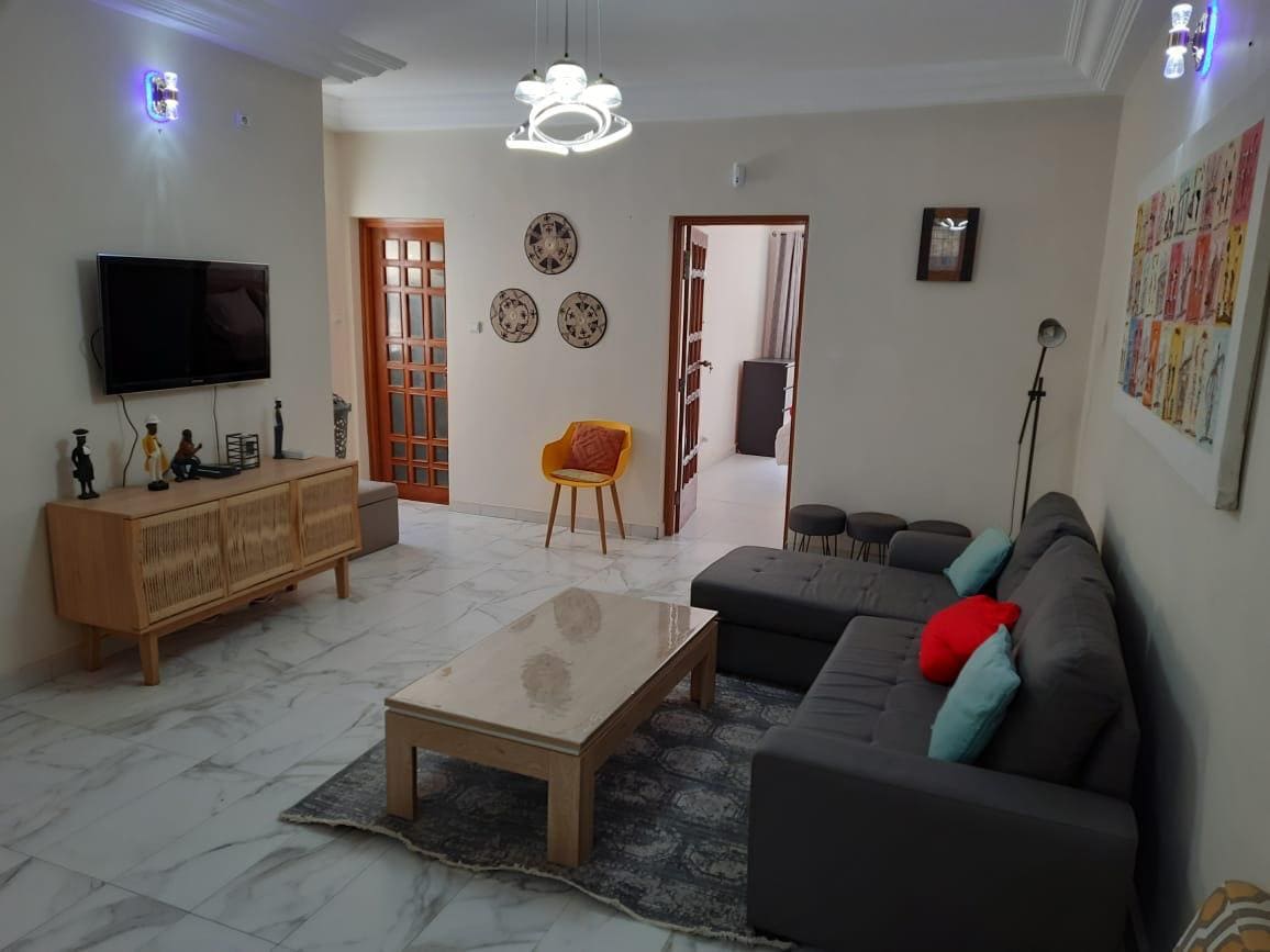 The living area features a modern gray sofa accompanied by colorful cushions, set against marble flooring. A wooden coffee table occupies the center, with a flat-screen television mounted on the wall. Warm lighting highlights the decorative wall art and wooden accents throughout the space.