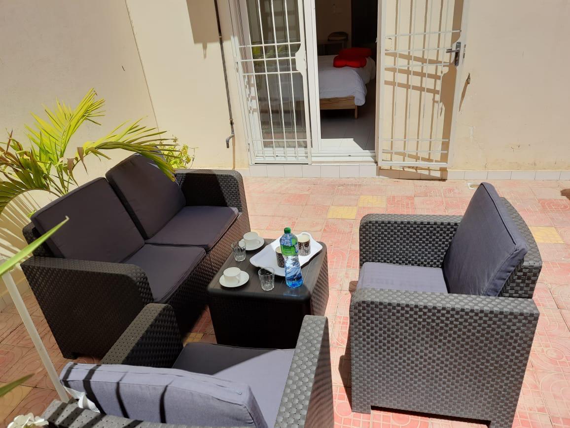 An outdoor seating area is depicted, featuring a set of four cushioned chairs surrounding a small table. Sunlight filters onto the terracotta-tiled floor, with a doorway visible in the background leading to a spacious bedroom.