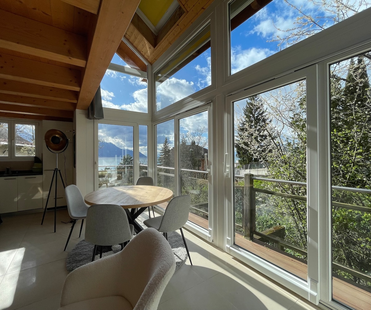A bright dining area is illuminated by natural light filtering through expansive glass windows. A round wooden table is surrounded by soft, upholstered chairs. Views of the lake and surrounding greenery are visible outside, creating a seamless connection with nature.
