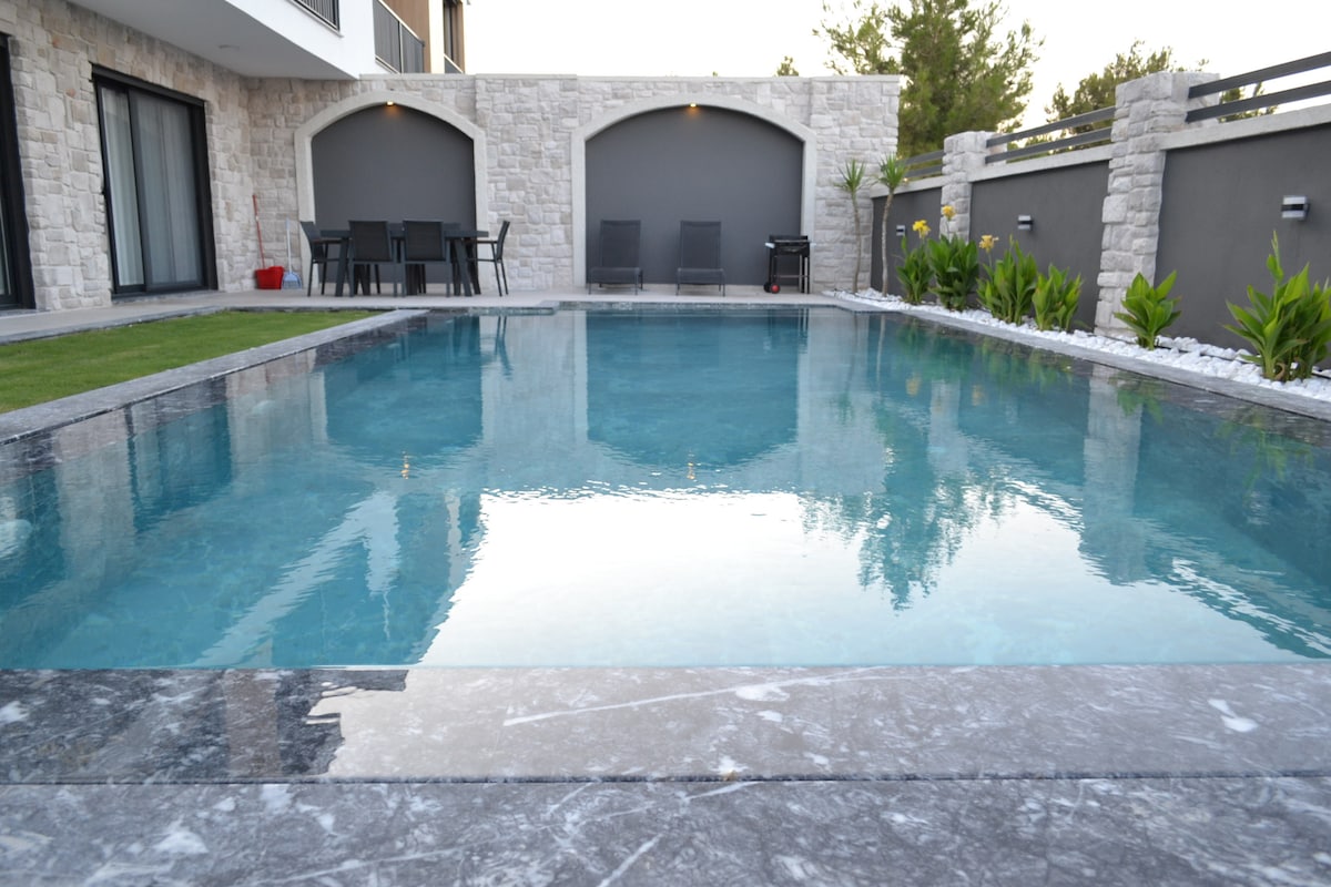 The image displays a tranquil private pool surrounded by natural stone walls and lush greenery. Smooth marble borders frame the clear water, reflecting the serene environment. Outdoor seating is visible in the background, offering a place for relaxation and enjoyment.