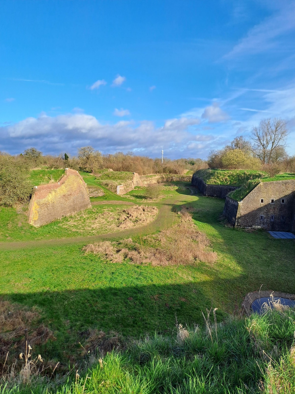 An expansive green landscape is viewed, featuring remnants of fortifications amidst rolling hills. Structures are partially covered in grass and surrounded by patches of trees. A clear blue sky enhances the serene setting, offering a glimpse of historical architecture in a natural environment.