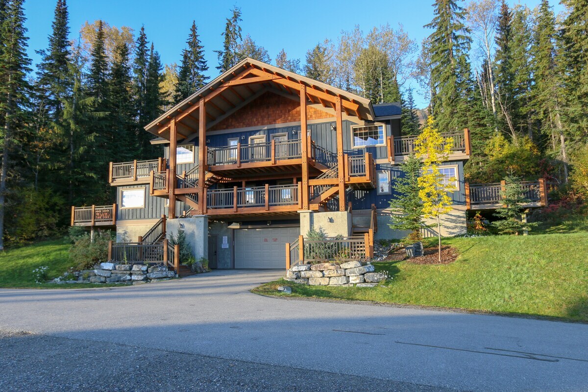 A multi-level building is nestled among tall evergreen trees, showcasing a rustic exterior with wooden beams and large balconies. A well-maintained lawn features scattered rocks and a landscaped area, providing a natural setting. A paved pathway leads toward the entrance from the street.
