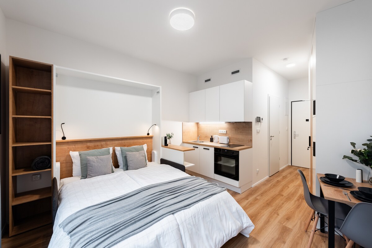 A modern studio features a spacious bed with gray and white bedding. A functional kitchenette is equipped with an oven, sink, and kettle, along with a dining area that includes a round table and two chairs. Ample natural light fills the room.