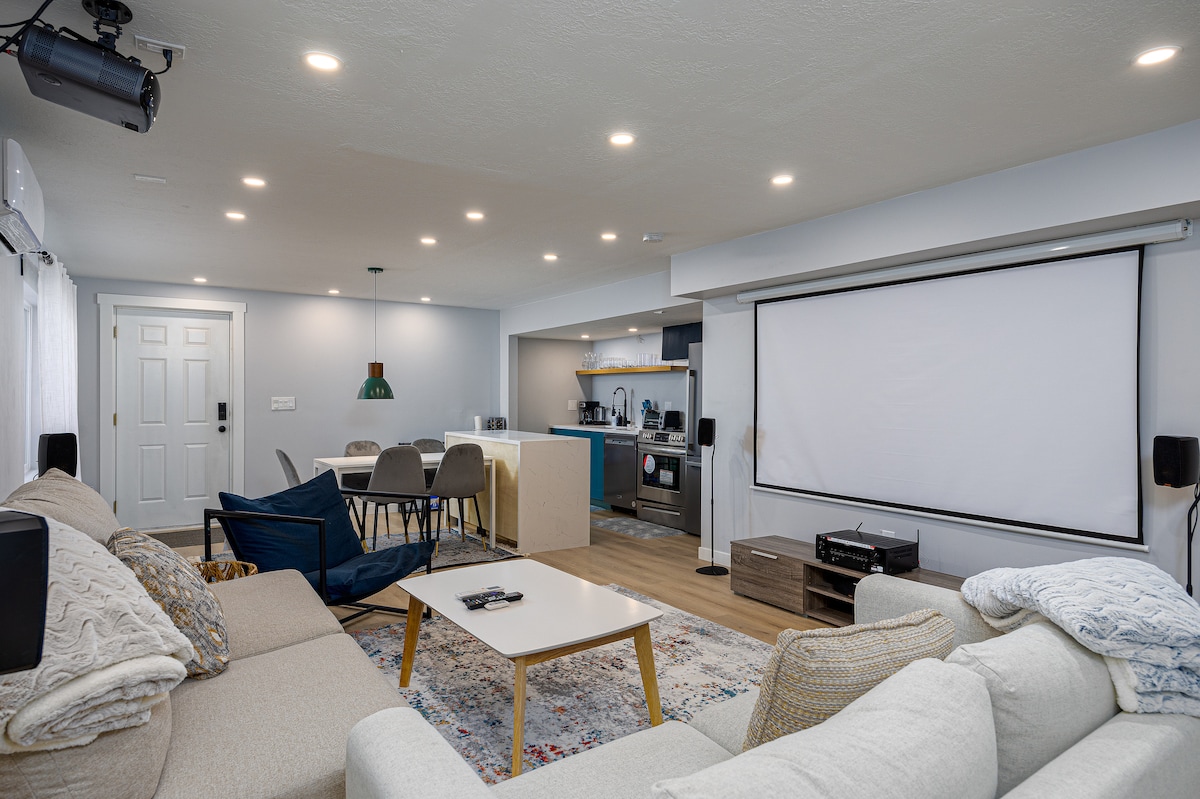 A cozy living space features a large white projection screen and comfortable seating arranged around a coffee table. A dining area with a modern table and chairs is visible, along with a well-equipped kitchen in the background. Soft lighting enhances the inviting atmosphere.