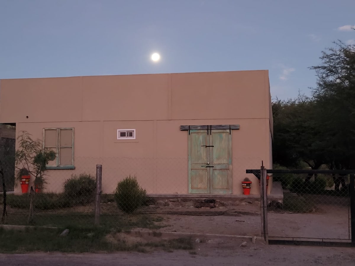 The exterior of the house is presented, featuring a soft sandy hue and rustic wooden doors. A full moon is visible in the twilight sky above, casting gentle light. A secure fence surrounds the property, complemented by greenery in the foreground.