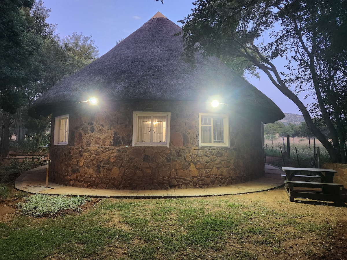 A round stone rondawel features a thatched roof and is surrounded by a landscaped area. Soft lighting illuminates the exterior, highlighting the natural textures of the stone and the inviting windowpanes. A picnic table is positioned nearby, enhancing outdoor seating options.