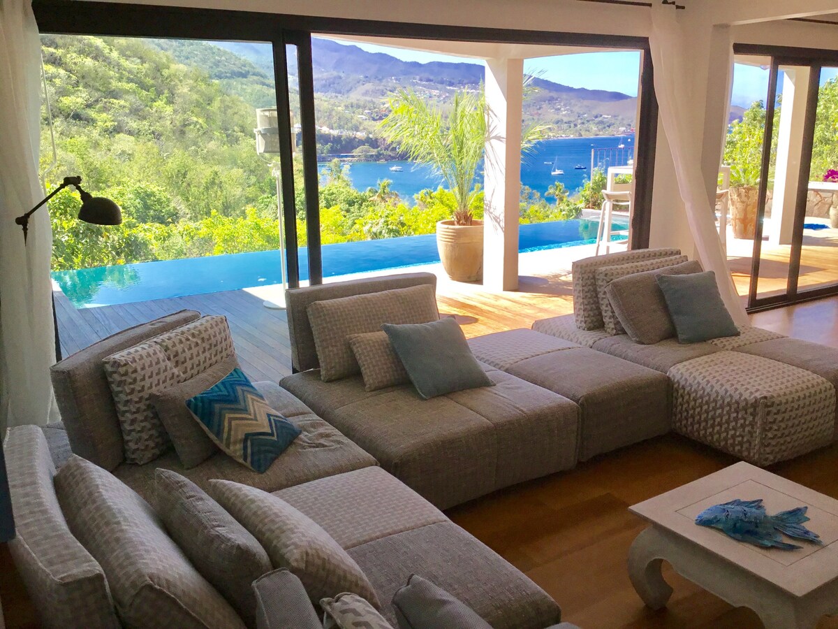 A spacious lounge area features a large sectional sofa arranged for relaxation, with light-colored fabric and decorative pillows. Expansive glass doors provide a view of a scenic outdoor area, showcasing greenery and a glimpse of a pool and distant hills.