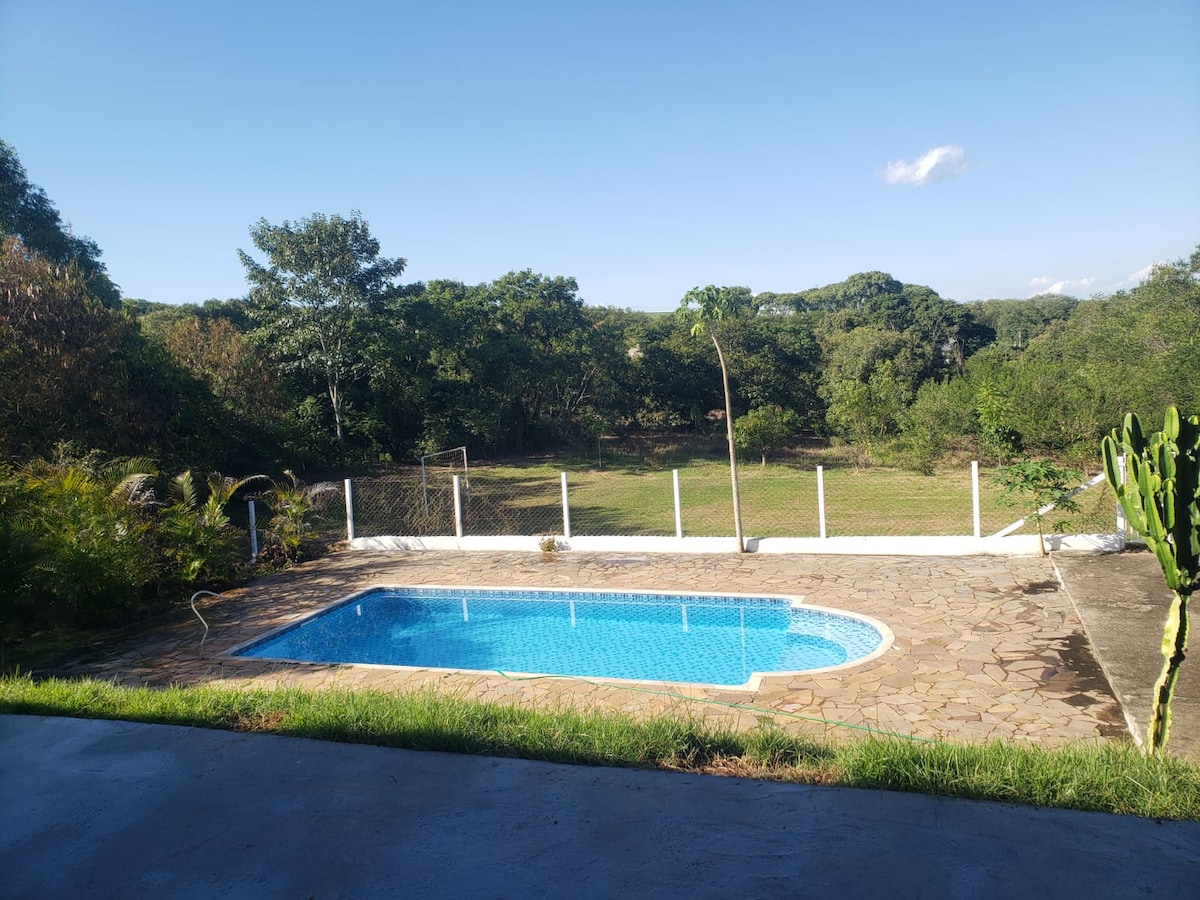 A serene outdoor space is displayed featuring a sparkling swimming pool surrounded by a stone patio. Lush greenery and trees create a natural backdrop, while a white fence outlines the area. The clear sky and soft sunlight enhance the tranquil atmosphere.