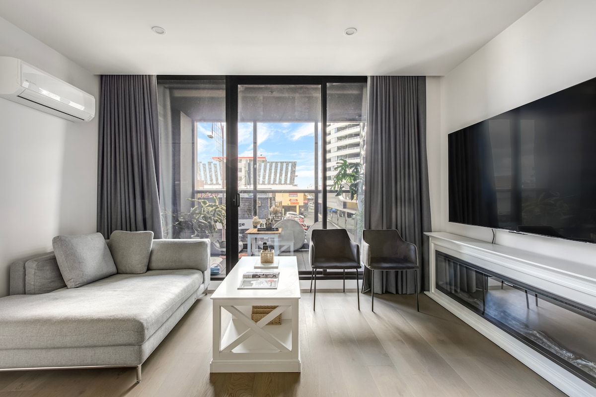 A modern living area features a cozy grey sectional sofa opposite a large flat-screen TV mounted on the wall. Floor-to-ceiling windows offer views of the vibrant surroundings, with natural light streaming in. A stylish coffee table sits in the center, complemented by two elegant chairs.