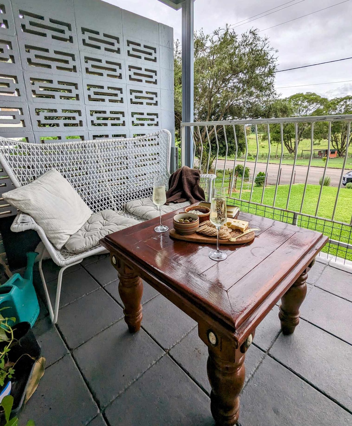 A cozy outdoor seating area features a woven chair and a wooden table. On the table, glasses and a small platter with snacks are arranged. The view includes lush greenery and a peaceful country road, enhancing a serene atmosphere.
