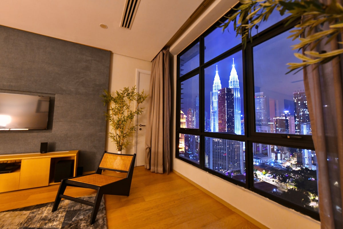 A modern living space features a large window showcasing illuminated views of the Petronas Twin Towers against a night sky. A sleek television is mounted on the wall, and a stylish chair complements the wood flooring. Soft drapes frame the window, enhancing the ambiance.