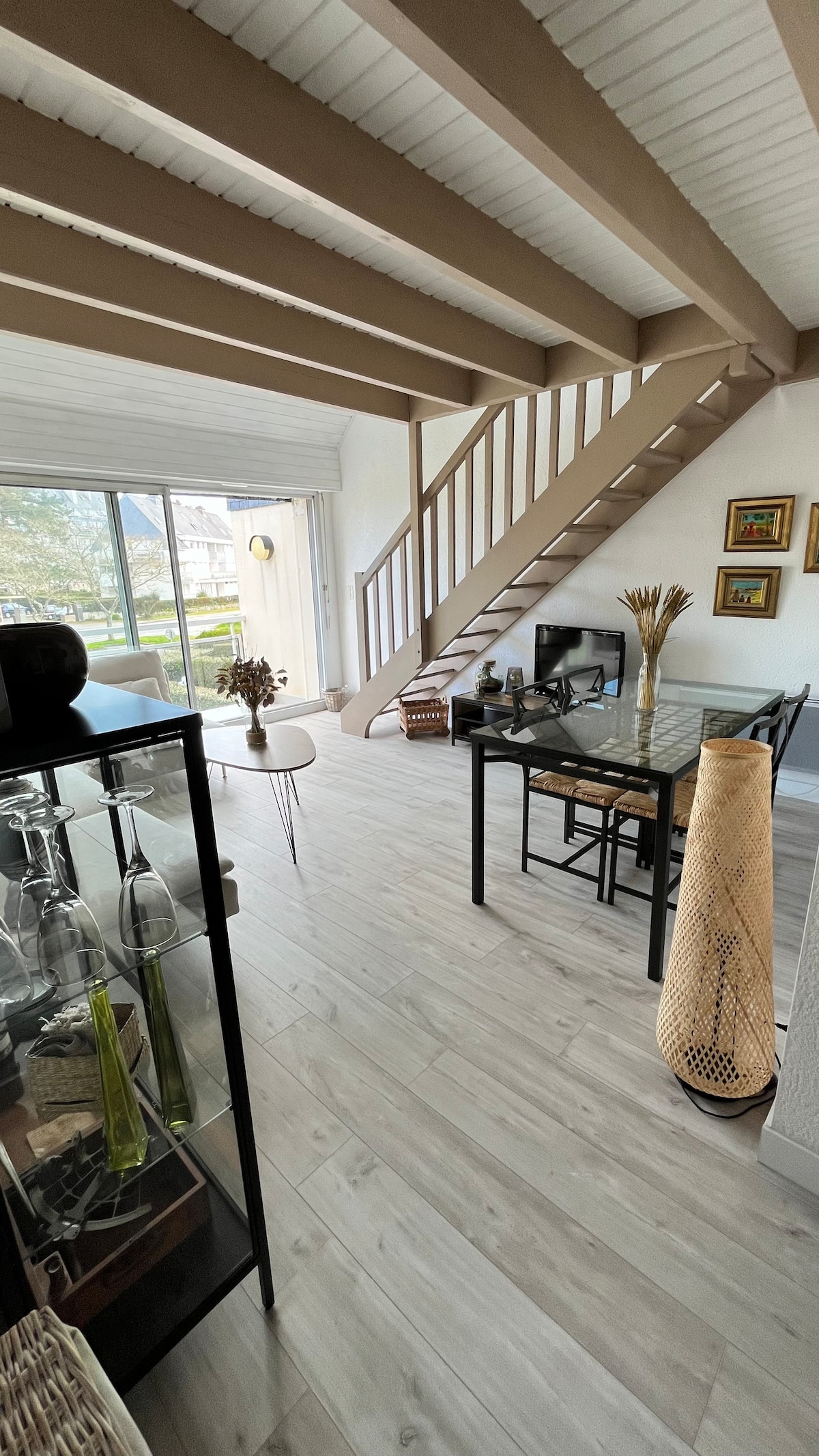 An open living area features light-colored wooden beams overhead, creating a sense of spaciousness. A glass dining table and white chairs are positioned near a large window, while a small staircase leads to the upper level. Decorative elements enhance the inviting environment.