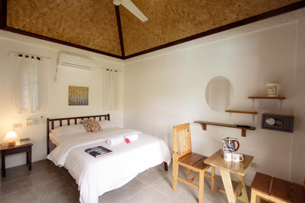 A well-lit bedroom features a spacious bed with a light-colored duvet arranged neatly. A wooden chair and a small table sit beside the bed. A mirror and a shelf are mounted on the wall, adding to the room's functionality. Natural light filters through the window treatments.