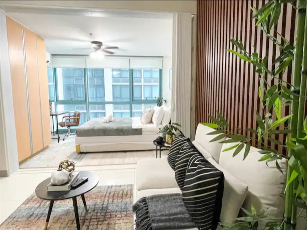 A modern living area features a light-colored sofa adorned with patterned pillows, positioned on a textured rug. A spacious sleeping area is visible in the background, separated by sliding glass doors, while a small table and chair are positioned by a large window.