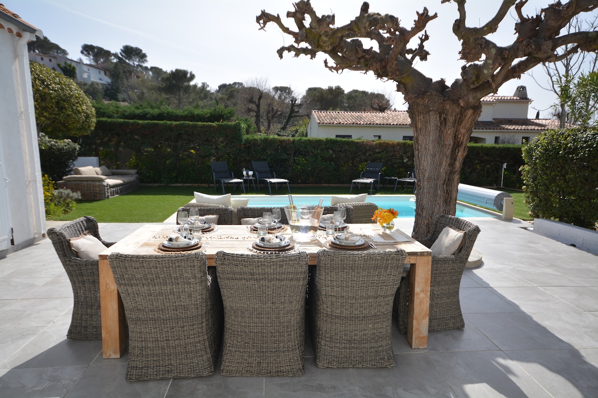 An outdoor dining area featuring a large wooden table surrounded by six wicker chairs. A blooming tree provides shade, and a swimming pool is visible in the background alongside manicured grass and lounging chairs.