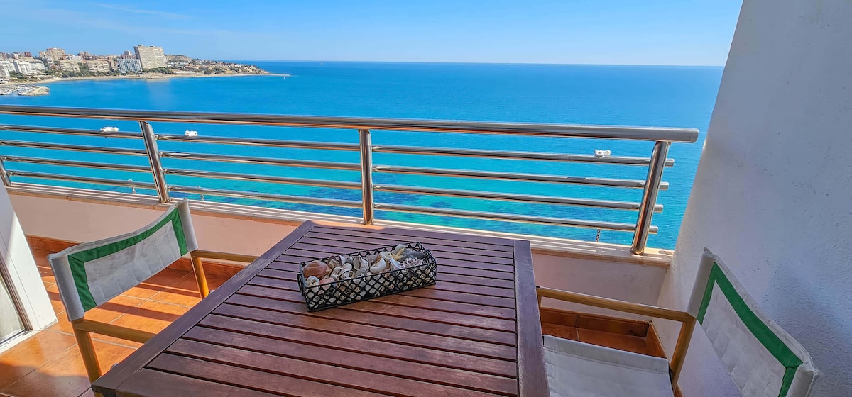 A terrace with a wooden dining table and two chairs offers breathtaking views of the sea. A decorative basket filled with seashells sits at the center of the table. The clear blue waters and distant coastline create a serene coastal backdrop.