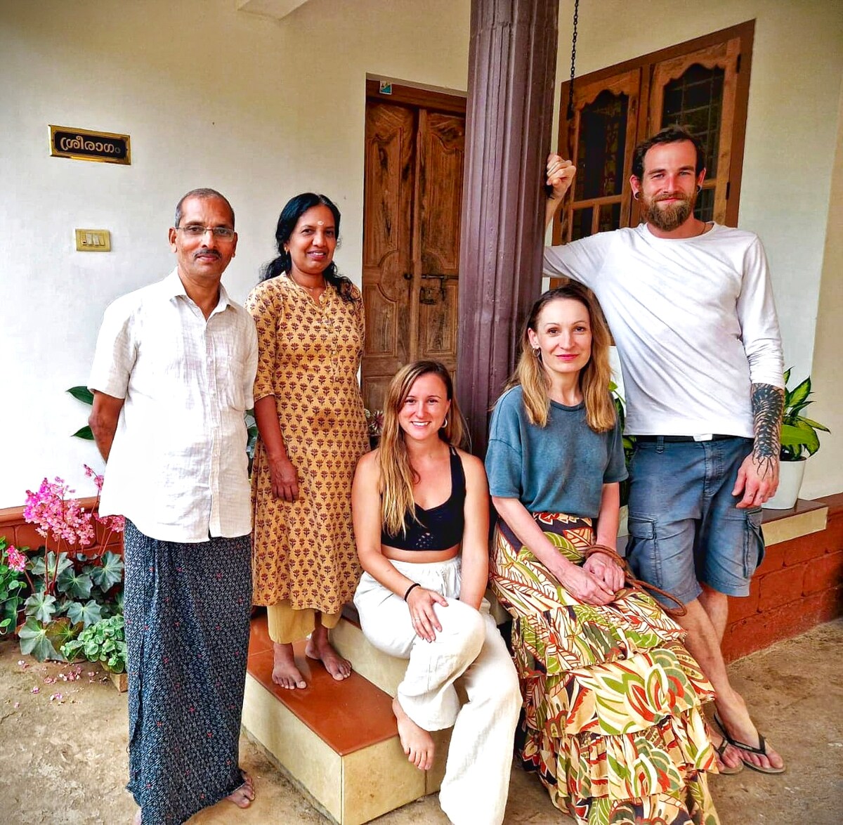 A group of five individuals is gathered outside a traditional building, characterized by wooden doors and decorative elements. Floral plants enhance the entrance area, while a wooden pillar supports an overhead swing. The surroundings reflect a warm and welcoming atmosphere.