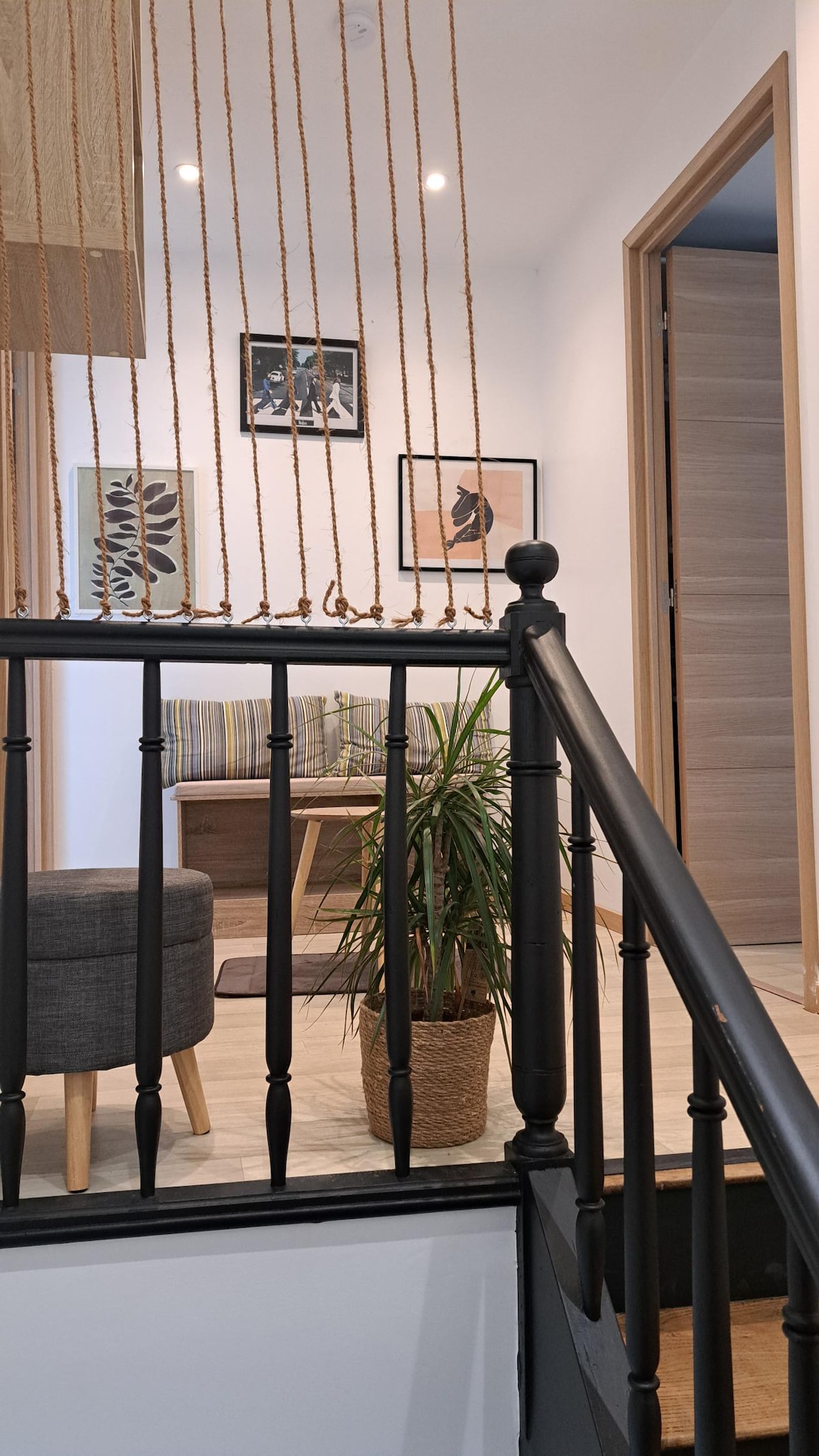 A view from the staircase showcases a cozy seating area with striped cushions and artwork on the wall. A potted plant adds a touch of greenery. The railing features vertical rope accents, leading toward a door that appears to open into another room.