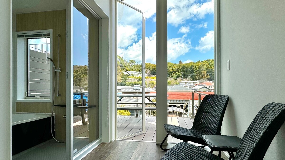 An open room features large glass doors that lead to a spacious balcony. Two black chairs are positioned indoors, offering a view of the surrounding greenery and rooftops. The modern interior is brightened by natural light streaming through the doors.