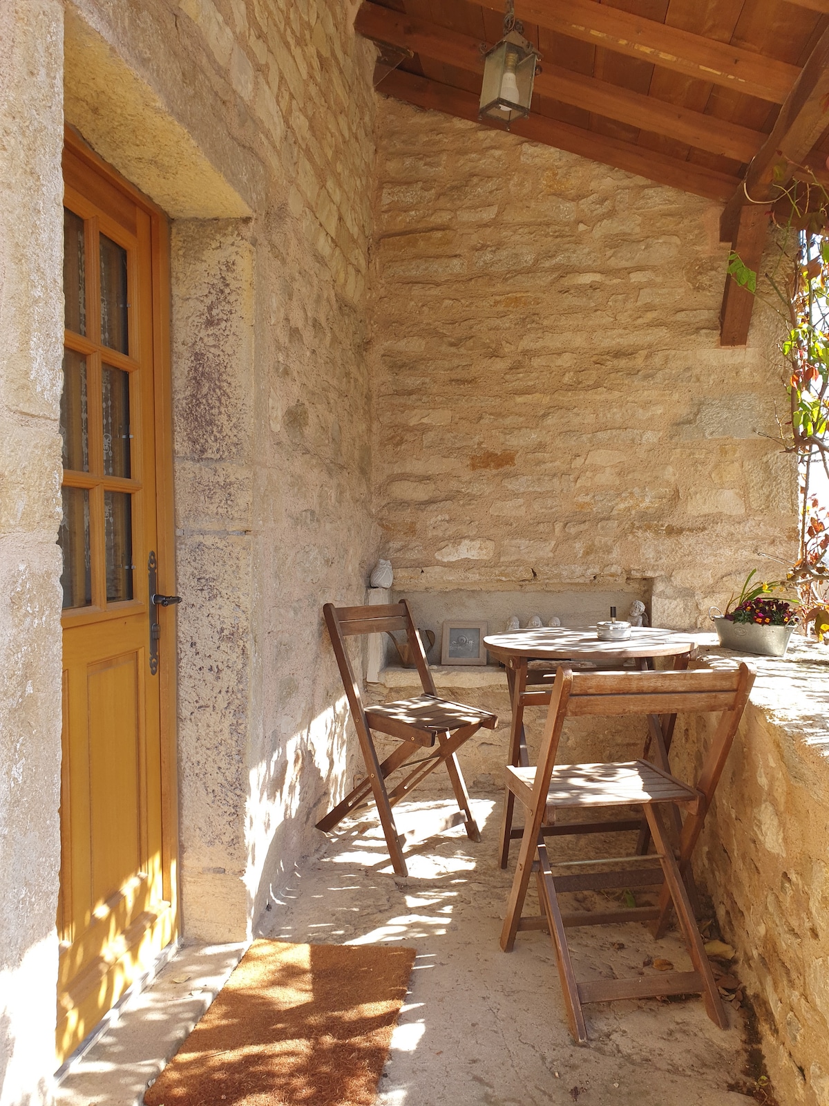 A small outdoor terrace is featured, showcasing a wooden table with two folding chairs. The stone walls reflect natural hues, and sunlight creates a warm ambiance. A potted plant adds a touch of greenery, while a door with a paneled design offers access to the interior.