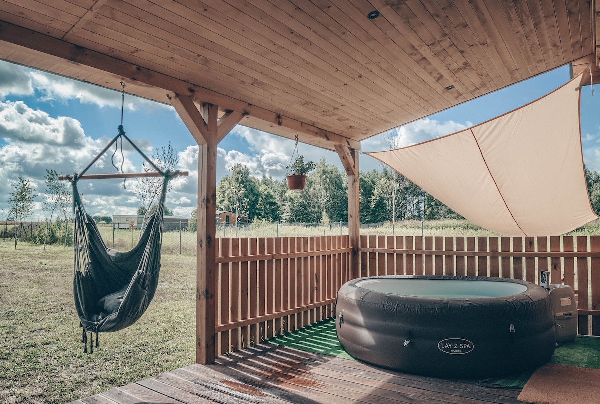 A covered terrace features a hanging chair and a heated jacuzzi. Natural light brightens the area, with cloud patterns visible in the sky. The terrace is surrounded by wooden fencing, creating a private outdoor space for relaxation.