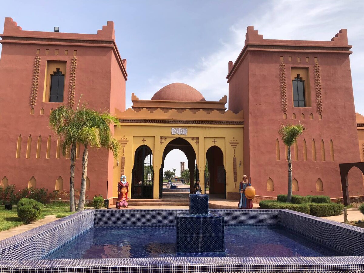 The entrance of the resort is framed by distinct architectural features in warm colors. A fountain is situated at the center, surrounded by a tiled pool. Lush palm trees and landscaped greenery complement the inviting courtyard area, enhancing the welcoming appearance.