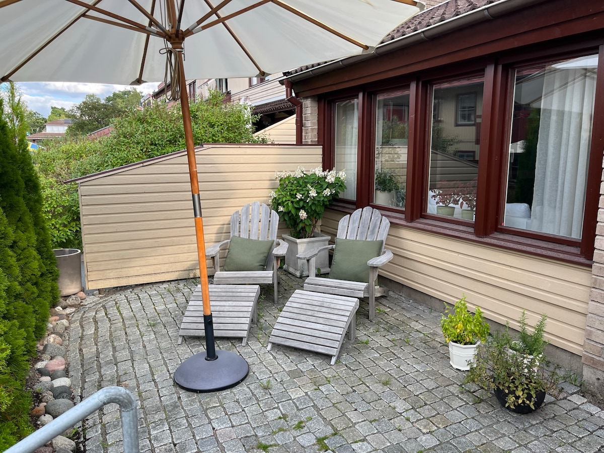 A shaded outdoor patio area is presented, featuring two wooden lounge chairs with cushions placed beside a small table. Decorative potted plants add greenery, while low stone walls provide privacy. Sunlight filters through an umbrella, creating a comfortable space for relaxation.