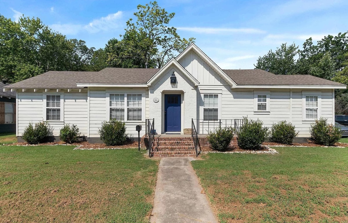 A charming single-story home is displayed with a welcoming blue front door. Steps lead to the entrance, bordered by manicured shrubs and a grassy yard. Large windows offer natural light, and a symmetrical design enhances the overall appeal.