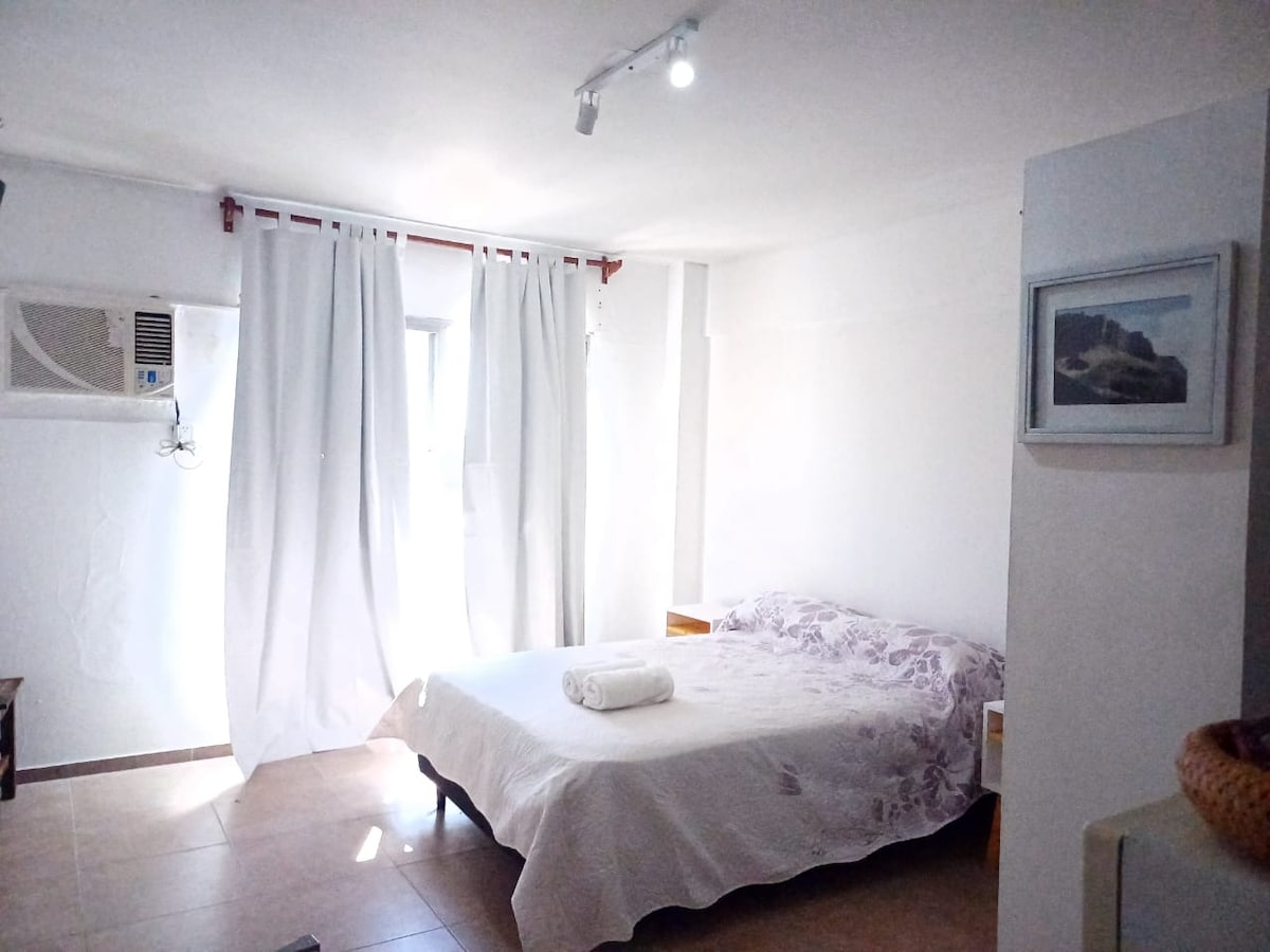 A well-lit bedroom features a spacious bed dressed in a white covering. Light curtains hang beside a window, allowing natural light to fill the room. A small side table is positioned next to the bed, and an air conditioning unit is mounted on the wall.