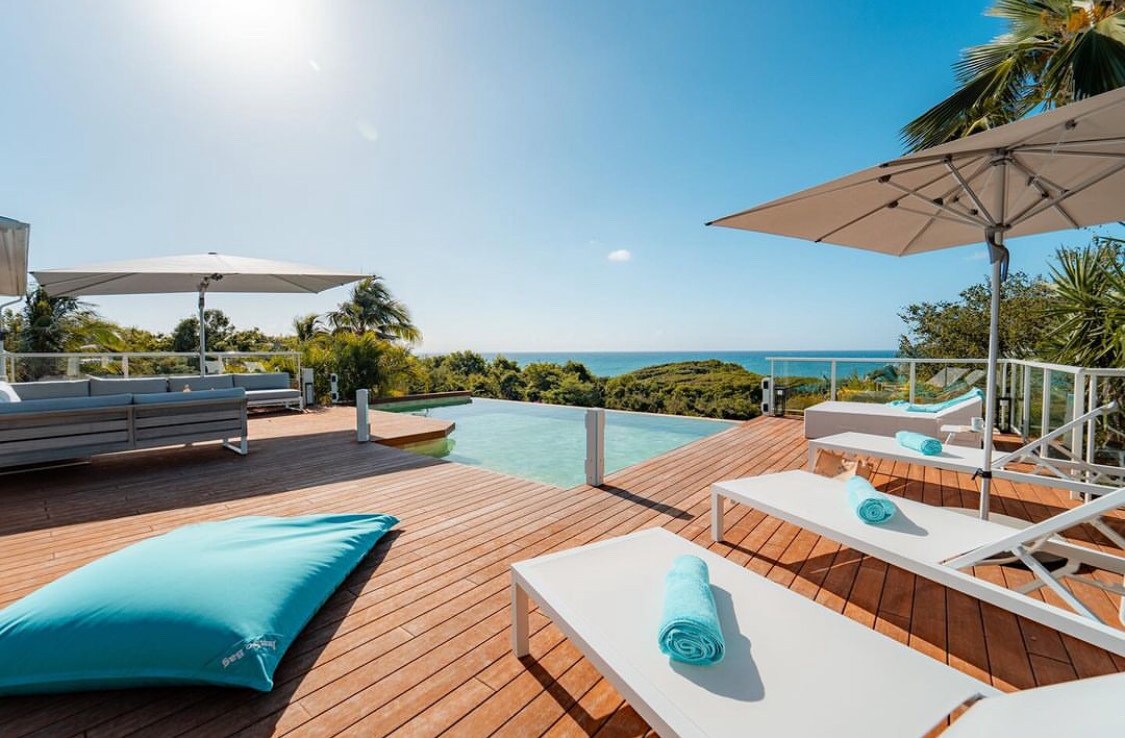 A spacious wooden deck surrounds an infinity pool, offering a panoramic sea view. Comfortable sun loungers and oversized cushions are positioned for relaxation, while large umbrellas provide shade from the sun. Lush greenery is visible in the background, enhancing the tranquil atmosphere.