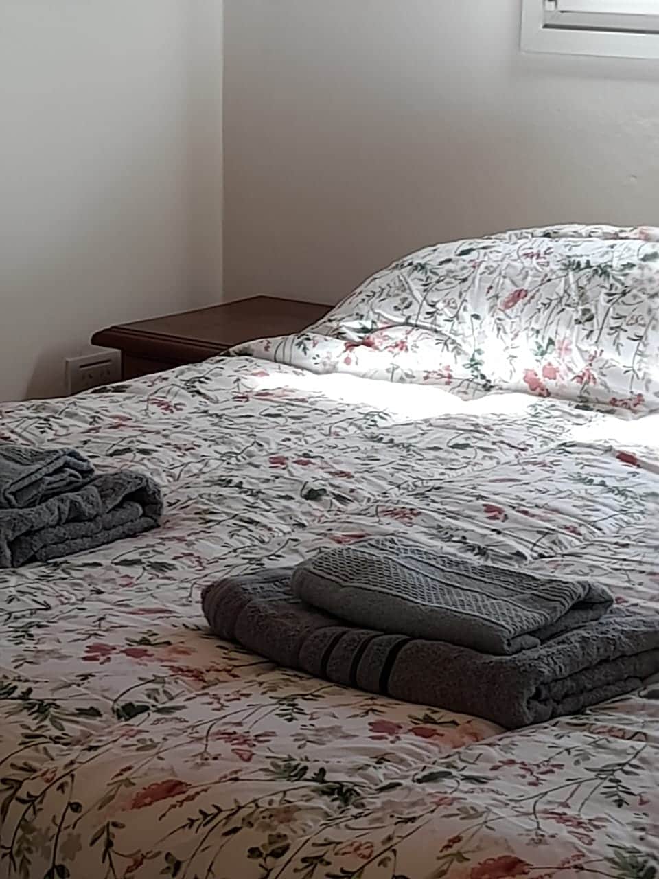 A neatly arranged sleeping area features a floral-patterned bedspread on a spacious bed. Folded towels are placed neatly at the foot of the bed and on a bedside table. Soft natural light filters in through a nearby window.