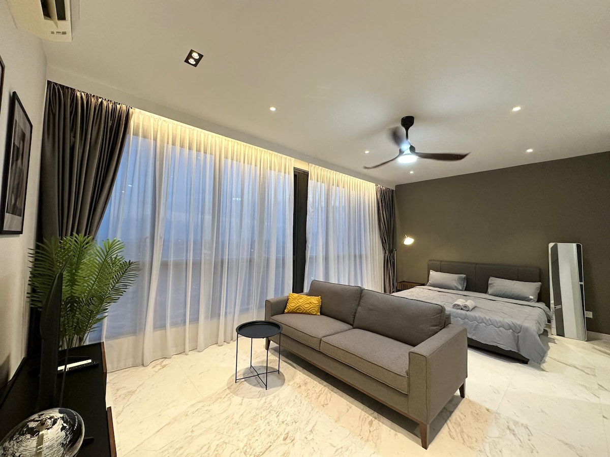 A spacious living area features a modern sofa, paired with a small round table. The large windows, dressed with floor-to-ceiling curtains, allow soft natural light to fill the room. A king-size bed is positioned in the background, complementing the neutral color scheme.