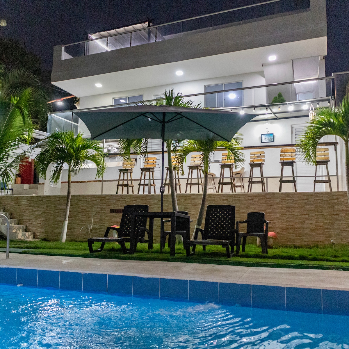 The image showcases a modern two-story house at night, with illuminated balconies and a seating area featuring bar stools. A large umbrella provides shade over a set of dark outdoor chairs near a vibrant pool, surrounded by lush greenery.