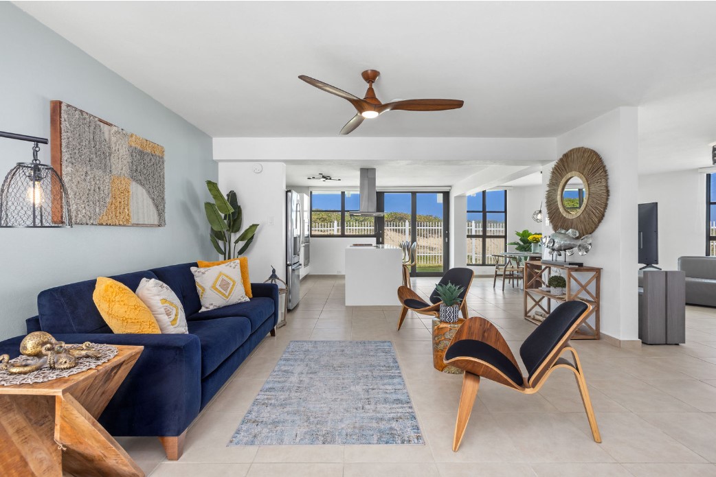 A spacious living area features a navy blue sofa adorned with yellow cushions. A wooden table holds decorative items, and unique chairs sit on either side of a textured area rug. Large windows provide natural light and views of the outdoor space.