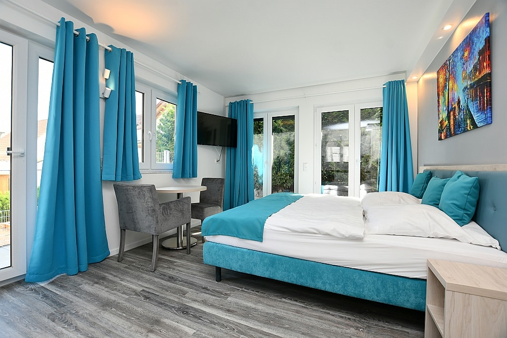 A modern sleeping area features a spacious bed dressed in crisp white linens with a teal accent headboard. Large windows allow natural light to fill the room, complemented by turquoise curtains. A small table and two chairs are positioned nearby, with a colorful artwork adorning the wall.