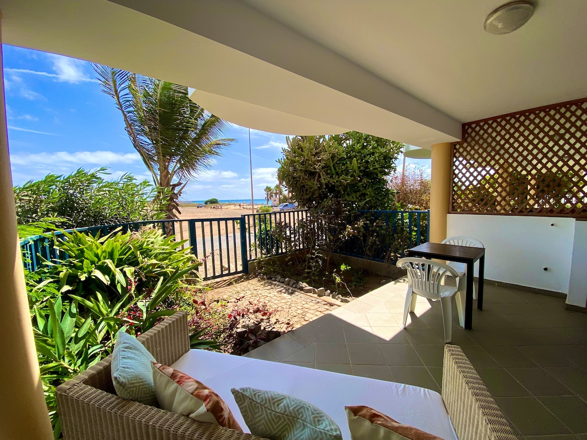 A terrace area is presented with a comfortable seating arrangement and a small table. Lush greenery borders the space, offering a view of the beach in the distance. The scene is brightened by natural light and features a clear blue sky.